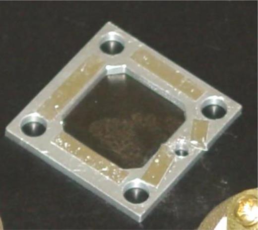 Handmade charge-control mesh-screen. 25 × 25 mm aluminum square (0.8 mm thick) with 90% transparent mesh taped down by hand using aluminum tape to one side of the square. Holes allow screws to be used if clips are not available. The mesh-screen is not perfectly flat. To produce the flat electric field, the aluminum needs to be grounded. Depending on the sample thickness, other mesh-screens were built using 1.0, 1.5, and 2.0 mm thicknesses.